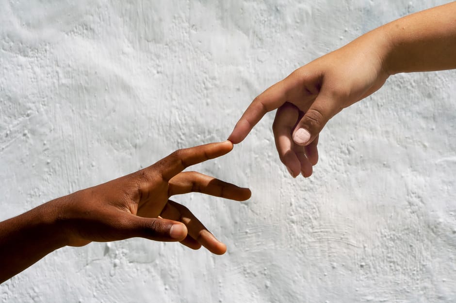 A close-up image depicting two human hands reaching towards each other against a textured white wall. One hand has darker skin tones and the other has lighter skin tones; each hand is extended with fi